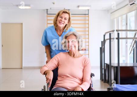 Una donna anziana siede su una sedia a rotelle con il suo fisioterapista in un centro di riabilitazione entrambi sorridenti. Questa immagine può essere utilizzata commercialmente per Healthca Foto Stock