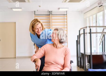 Una donna anziana su una sedia a rotelle sorride al suo fisioterapista in un centro di riabilitazione. Questa immagine può essere utilizzata in commercio per rappresentare il manuale di riabilitazione Foto Stock