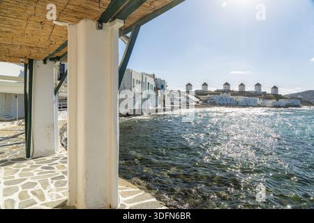 La vista degli edifici imbiancati incontra il mare turchese, con gli storici mulini a vento che si stagliano contro lo skyline sotto un sole luminoso, Mykonos, la città vecchia, G. Foto Stock