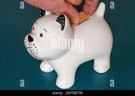 Primo piano di una mano che inserisce una moneta in euro in un salvadanaio in ceramica, simboleggiando risparmi, finanza, bilancio ed economia personale Foto Stock