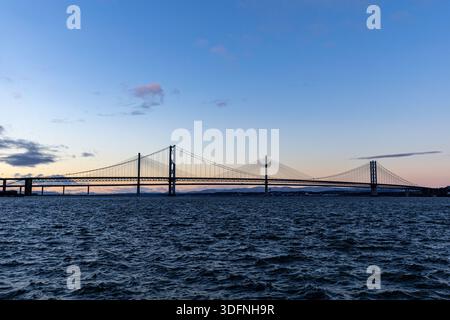 Un lungo ponte sospeso attraversa un corpo d'acqua sotto un cielo in pendenza di blu e arancione pallido al crepuscolo. Foto Stock