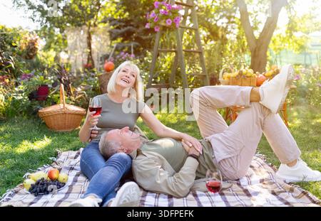 Romantico sposi anziani godersi un picnic insieme, riposare su coperta in giardino e bere vino rosso, spazio libero Foto Stock