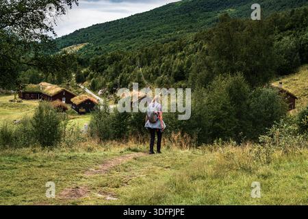 Una donna in spalla cammina lungo un sentiero escursionistico nella valle di Innerdalen. Le tradizionali cabine norvegesi in legno sul tetto sono visibili tra gli alberi verdi delle colline Foto Stock