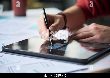 Primo piano di un uomo anonimo che utilizza lo stilo disegnando su un tablet digitale, appoggiando le mani sullo schermo, lavorando al progetto con documenti e schizzi visibili sulla scrivania in background Foto Stock