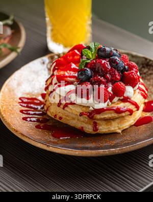 Una deliziosa pila di soffici pancake conditi con fragole fresche, mirtilli, lamponi, panna montata e sciroppo di frutta rossa. Foto Stock