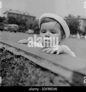 Una fotografia d'archivio in bianco e nero di Slaviansk, URSS, scattata negli anni '1960, presenta un ritratto ravvicinato della bambina Inessa Mikhailovna Kravelis. Indossando un berretto bianco, si appoggia su un confine di cemento in Piazza della Rivoluzione d'ottobre (ora Piazza Soborna), potendo le mani sul bordo. Guarda avanti con un'espressione penosa e seria, le labbra si sono leggermente staccate. Lo sfondo sfocato mostra veicoli sovietici d'epoca ed edifici cittadini. Questa immagine candida cattura un momento tranquillo e contemplativo dell'infanzia nella pacifica regione del Donbas. Foto Stock