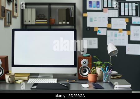 Spazio di lavoro moderno con un grande computer desktop con schermo vuoto sulla scrivania, tablet da disegno digitale, altoparlanti, piante in vaso, forniture per ufficio, scaffalature e disegni a parete Foto Stock