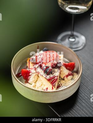 Una deliziosa fetta di cremosa cheesecake basca bruciata servita in un recipiente di ceramica, condita con fragole fresche, mirtilli e zucchero a velo. Foto Stock