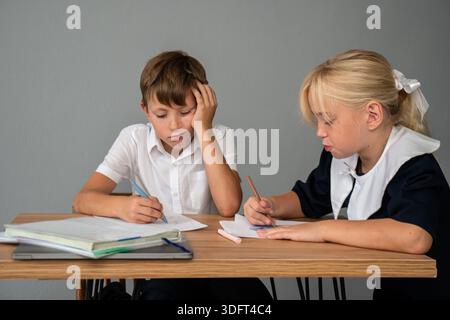 Studio dei compiti scolastici per bambini - due bambini in uniforme scolastica scrivono in quaderni a una scrivania, uno sembra stanco. Foto Stock