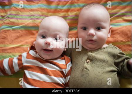 Due felici fratelli gemelli giacciono fianco a fianco su una coperta colorata a righe. Un bambino sorride mentre l'altro sembra curioso. Sono molto vicini e.. Foto Stock