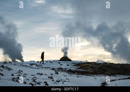 Vista di una figura solitaria che attraversa il paesaggio aspro e innevato tra i pennacchi di vapore geotermico che si innalzano dalla terra, Rejkjahlid, Icela settentrionale Foto Stock