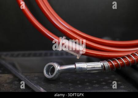 Tubo flessibile della tubazione freno intrecciato rosso resistente e durevole con raccordo maschio in metallo e collegamento in acciaio che posa in garage con superficie ruvida scura che rappresenta l'industria Foto Stock