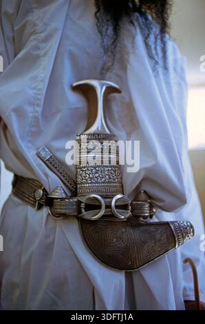 Detailaufnahme eines kunstvoll verzierten traditionellen Dolchs mit gebogener Klinge in einer reich ornamentierten Scheide. Der Dolch ist am Gürtel eines Trägers befestigt und Teil traditioneller Kleidung. Oman, mai 1975. Foto Stock