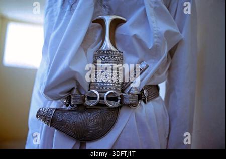 Detailaufnahme eines kunstvoll verzierten traditionellen Dolchs mit gebogener Klinge in einer reich ornamentierten Scheide. Der Dolch ist am Gürtel eines Trägers befestigt und Teil traditioneller Kleidung. Oman, mai 1975. Foto Stock