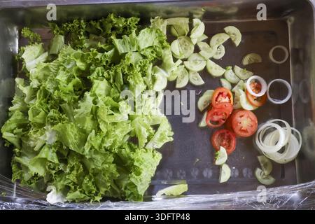 Insalata di lattuga verde fresca tritata con cetriolo a fette di pomodoro anello di cipolla vassoio di metallo preparato ricoperto di involucro di plastica per una dieta sana cottura dei pasti Foto Stock
