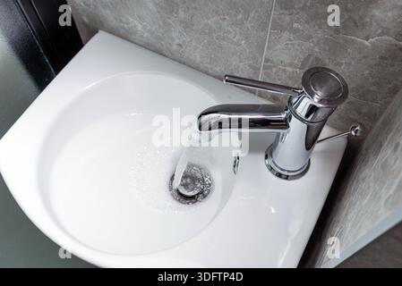 Rubinetto d'acqua cromato dal design moderno che scorre in un lavello in ceramica bianca. Vista dall'alto. Foto Stock