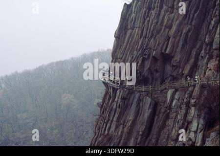 Passerella scavata nella parete rocciosa del monte Songshan, con i visitatori che si spostano lungo uno stretto sentiero sopra una valle nebbiosa e boscosa Foto Stock