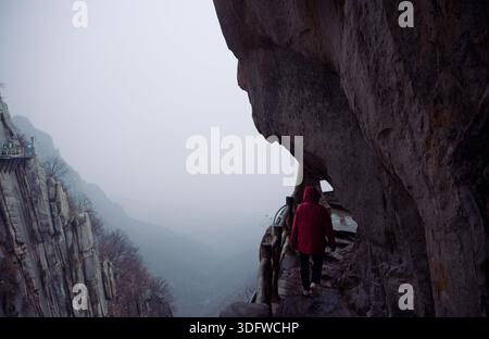 L'escursionista con una giacca rossa cammina lungo uno stretto sentiero sulla scogliera scavato nella parete rocciosa del monte Songshan Foto Stock