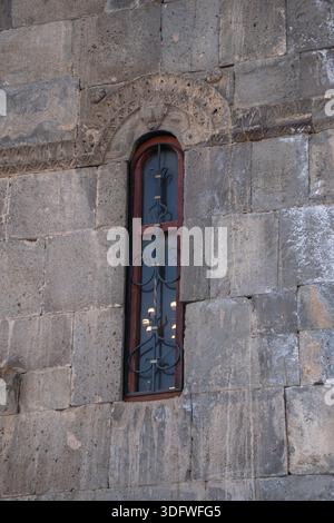 Stretta finestra ad arco con griglia in metallo adagiata su pareti in pietra del Monastero di Tatev, blocchi di pietra calcarea grigia intemprati, luce soffusa, calma atmosfera storica Foto Stock