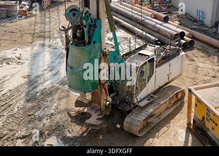 Un grande pezzo di attrezzatura da costruzione sta perforando attivamente il terreno in un cantiere affollato, riempito di tubi e attrezzi. Foto Stock