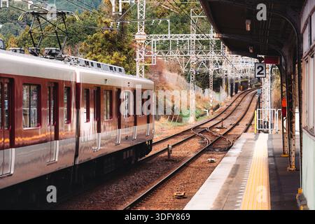 Treno locale con carrozze rosse e bianche che lascia la stazione ferroviaria rurale con binari, linee elettriche e pali elettrici in Giappone Foto Stock