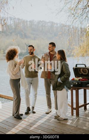Quattro amici si riuniscono su un ponte di legno vicino all'acqua, condividendo bevande e risate mentre si godono la splendida giornata autunnale. Foto Stock