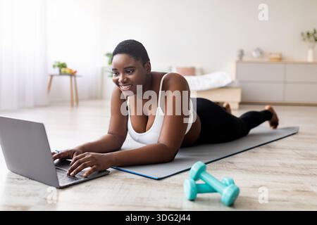 Donna nera curvy che guarda video sportivi online su un laptop, sdraiata a terra e che usa gadget prima di allenarsi in casa Foto Stock