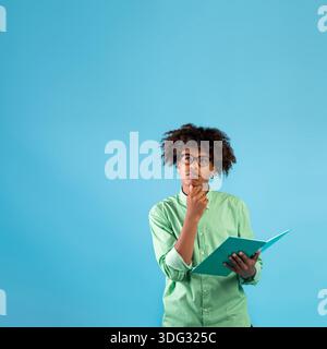 Premuroso ragazzo teen afro-americano in occhiali che tengono il notebook e brocciare il mento, guardando in su allo spazio libero, raccolto Foto Stock