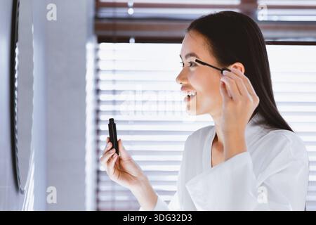 Donna applica Mascara mentre guarda in Mirror durante la routine mattutina a casa Foto Stock