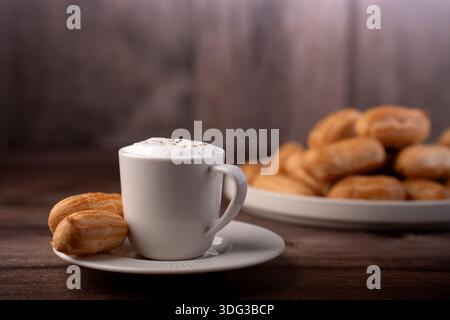 primo piano di cappuccino schiumoso in una tazza di ceramica bianca su un piattino con schiuma cosparsa di cacao, accanto a un piatto ricoperto di pasticcini dorati con panna e spugne su un Foto Stock