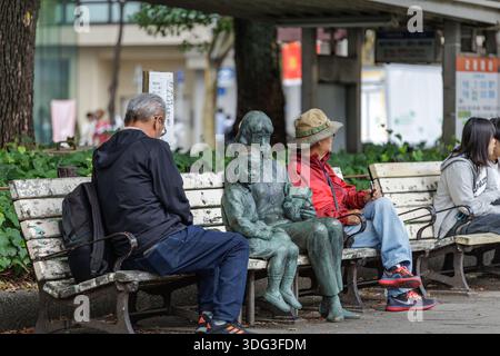 La statua di bronzo di una donna e di un bambino siede tra i parkgoers su una panchina intemprata in un parco urbano giapponese, giustapponendo l'arte pubblica alla vita quotidiana. Foto Stock