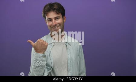 L'uomo punta il pollice su un lato in uno studio viola indossando una camicia in denim chiaro e una T-shirt bianca, sorridendo mentre guardi fuori dalla fotocamera; informale e cordiale. Foto Stock