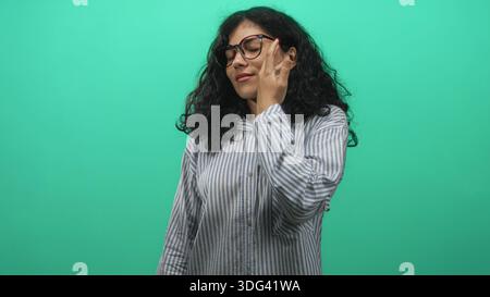 Giovane donna araba con capelli ricci e occhiali che toccano la guancia con mano in studio verde che indossa camicia a righe e colletto abbottonato; calma confida Foto Stock