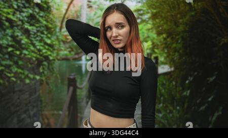 Giovane donna con la mano dietro il collo, sgombra esposta in piedi da una ringhiera di corda su una stretta passerella forestale accanto a un canale; disagio. Foto Stock