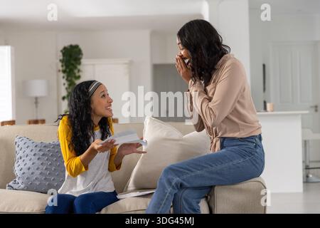 Mamma e figlia adolescente diverse sedute sul divano grigio leggono il biglietto d'auguri a casa con la tazza Foto Stock