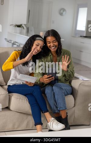 Mamma e figlia adolescente variegata che sventolano allo smartphone mentre tengono le lettere sul divano Foto Stock