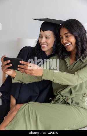 Mamma e figlia adolescente diverse che si fanno selfie sul divano con un abito da laurea e una tuta Foto Stock