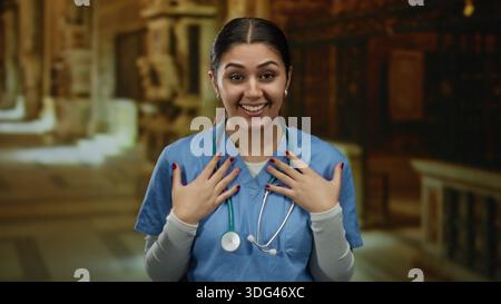 La giovane infermiera ispanica in scrub blu sorride in un ambiente di chiesa, trasmettendo calore e professionalità in un ambiente interno. Foto Stock