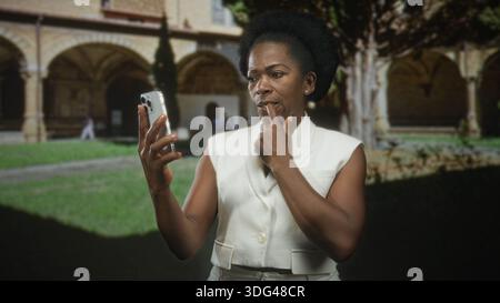 La donna tiene lo smartphone e le dita contro le labbra in posa con un giubbotto bianco e un afro, in piedi vicino agli archi dell'edificio; riflesso riflesso riflesso. Foto Stock