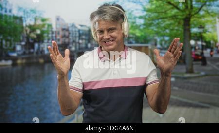 L'uomo con i capelli grigi che indossa le cuffie sulle orecchie sorride e regala due pollici mentre si trova in piedi accanto a un passaggio pedonale sul canale in una strada; felicità e svago. Foto Stock