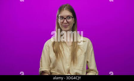 Giovane donna bionda che indossa una camicia beige che tiene il cuore rosso con entrambe le mani e sorride in un vivace studio viola; affetto. Foto Stock