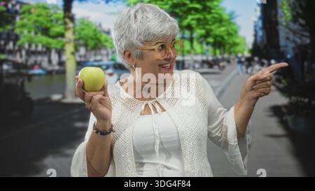 Donna che tiene la mela verde in mano sinistra e punta con la mano destra in strada, sorride con i capelli grigi e indossa gli occhiali; gioiosa curiosità. Foto Stock