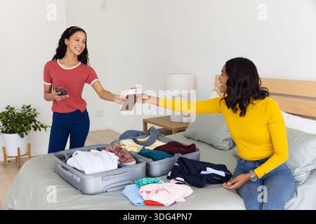 Mamma e figlia adolescente diverse sul letto scambiando passaporto e biglietto con il telefono vicino alla valigia Foto Stock