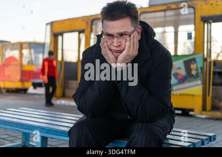 un uomo si siede alla fermata dell'autobus in attesa di un autobus, un tram, un tram. Viaggiare sui mezzi pubblici per andare al lavoro, studiare, all'università. In ritardo. Programma del percorso. Foto Stock