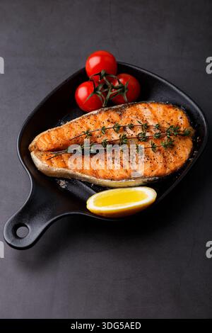 Salmone alla griglia con pomodori ciliegini, timo e limone su una piccola teglia nera. Il pesce fritto è un piatto tradizionale della cucina casalinga e dei ristoranti. Bla Foto Stock