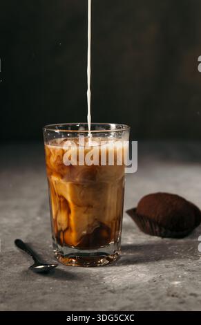 Il latte viene versato in un bicchiere di caffè ghiacciato con un dessert al cioccolato su sfondo scuro Foto Stock