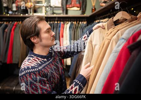 Bell'uomo consumatore che sceglie abbigliamento in boutique vintage di seconda mano, che sceglie la giacca da appendiabiti. Foto Stock