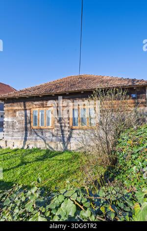 Un piccolo, vecchio cottage in legno si trova accanto ad una falsa recinzione in cemento, con edera e arbusti sovrastati. Le case moderne si affacciano su un quartiere tranquillo, sotto un br Foto Stock