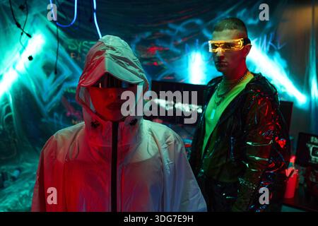Due giovani adulti, donna e uomo caucasici, con giacche futuristiche e occhiali visiera, in piedi in una stanza illuminata al neon con monitor per computer e sfondo graffiti, che incarna il tema hacker Foto Stock