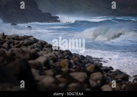 Le potenti onde dell'Atlantico si infrangono contro una spiaggia rocciosa di ciottoli sull'isola di Madeira, in Portogallo, con spruzzi e schiuma bianca che si snodano verso la costa sotto la luce del giorno Foto Stock